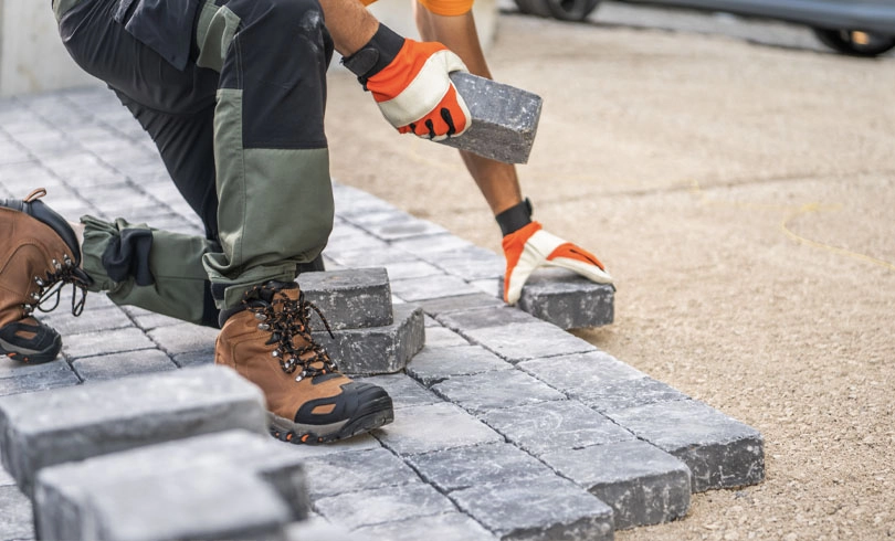 A construction worker is kneeling on the ground, carefully placing paving stones in a driveway. Wearing gloves and sturdy boots, they focus on arranging the stones