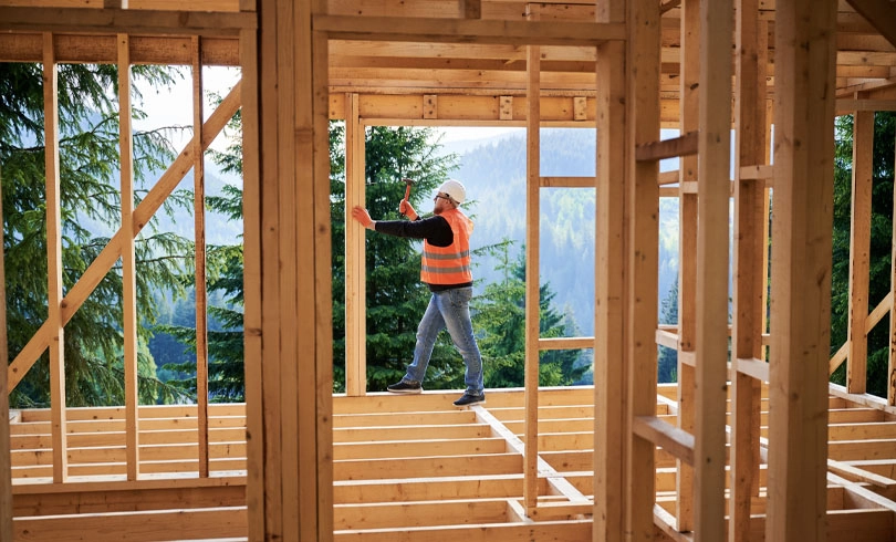 Carpenter constructing two-story wooden frame house near forest. Man hammering nails into the structure, wearing protective helmet and construction vest. Concept of modern ecological construction.